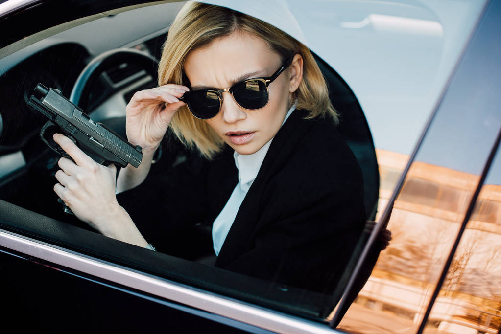 komoly fiatal szőke nő megható napszemüveg és gazdaság pisztoly az autóban  - Fotó, kép