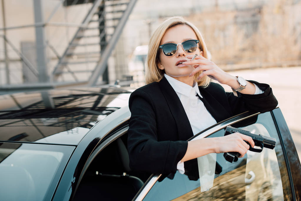 mujer rubia en gafas de sol fumar cigarrillo y pistola de mano cerca del coche
  - Foto, Imagen