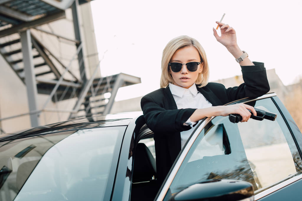 Piękna Blondynka palacz w okulary przeciwsłoneczne trzymając papierosa i pistolet w pobliżu samochodów  - Zdjęcie, obraz