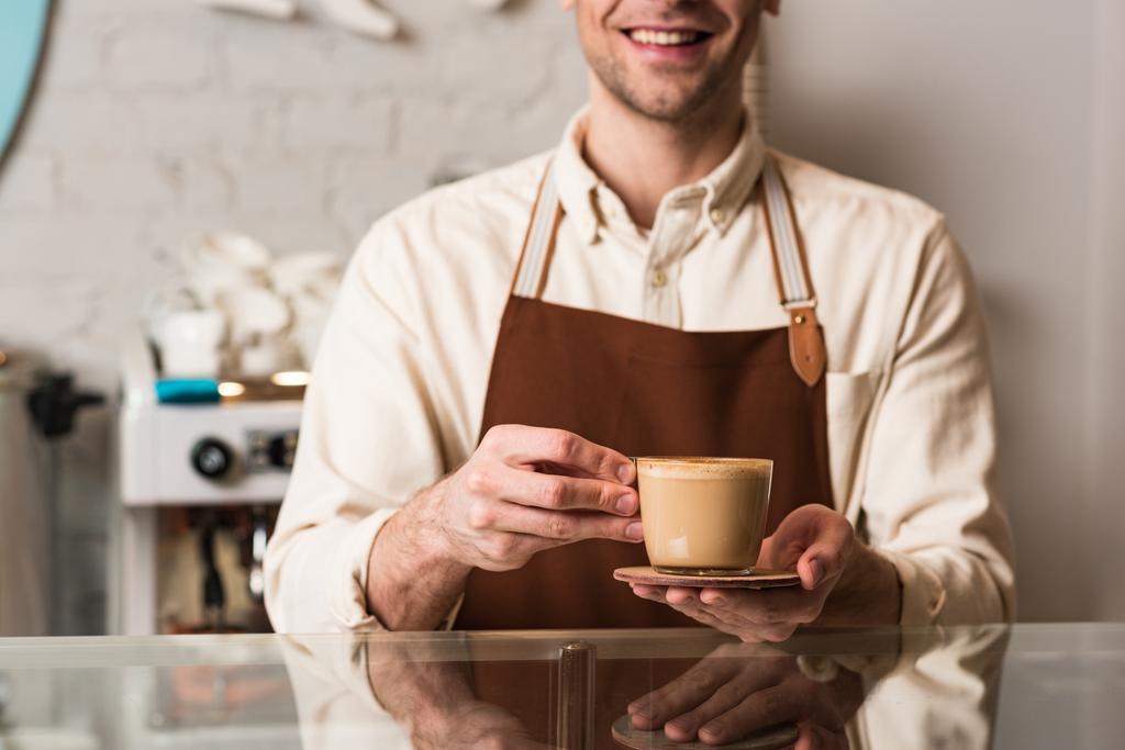 Обрезанный вид на баристу в коричневом фартуке с чашкой кофе с улыбкой
 - Фото, изображение
