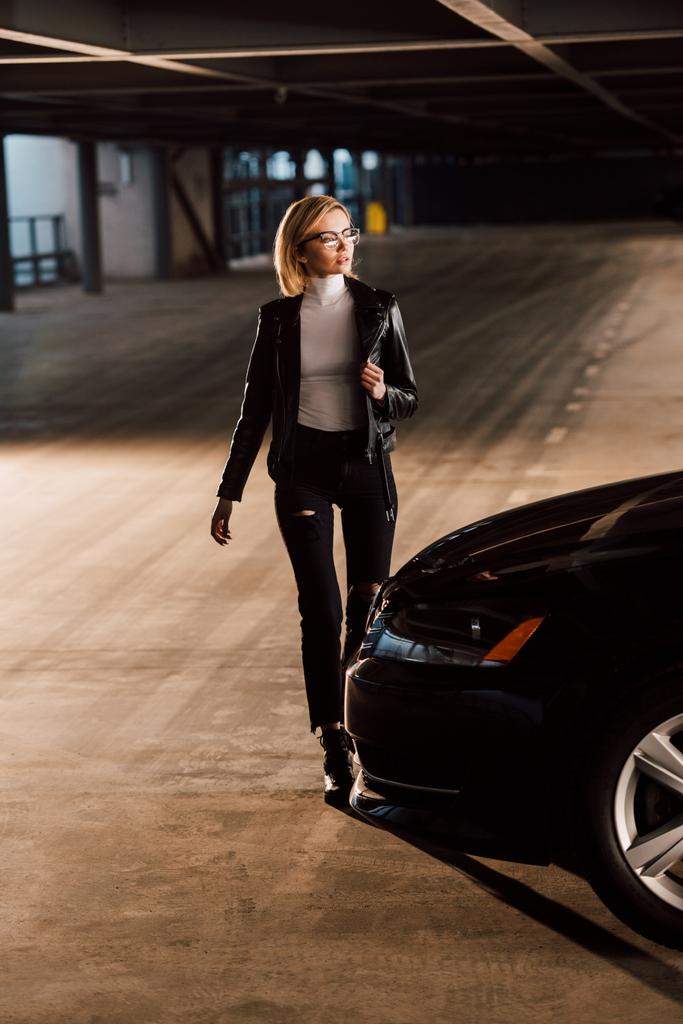 Hübsche junge blonde Frau mit Brille läuft auf Parkplatz neben Auto - Foto, Bild