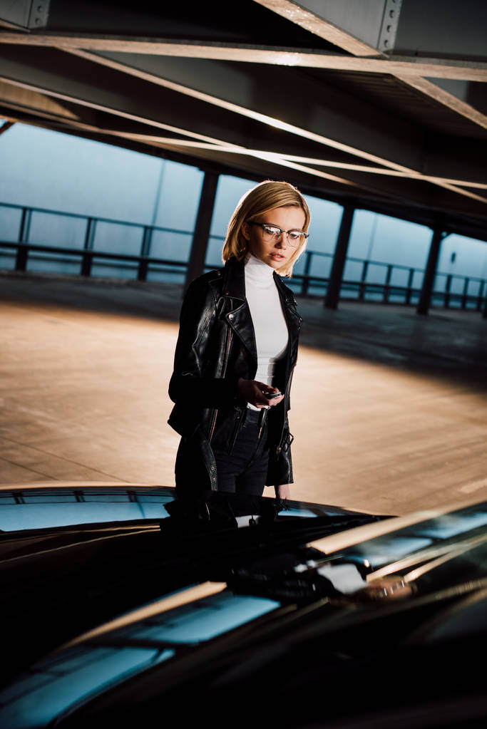atraente jovem mulher em óculos de pé no estacionamento perto de carro preto
 - Foto, Imagem