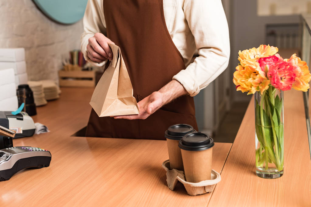 Partial view of barista in brown apron with paper bag and coffee to go - Photo, Image