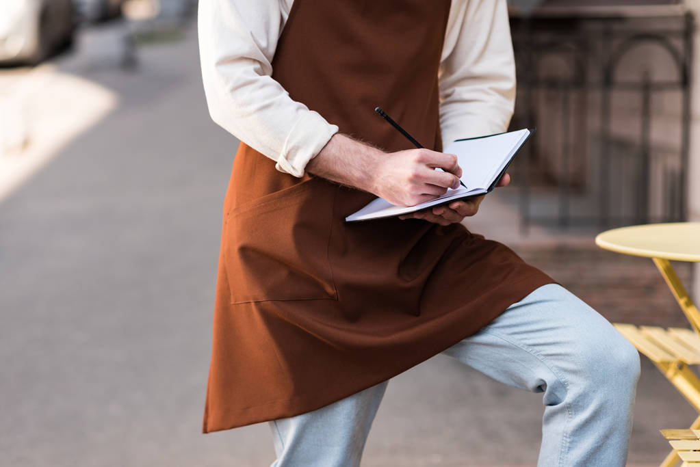 Обрезанный вид баристы в коричневом фартуке, пишущей в блокноте на улице
 - Фото, изображение