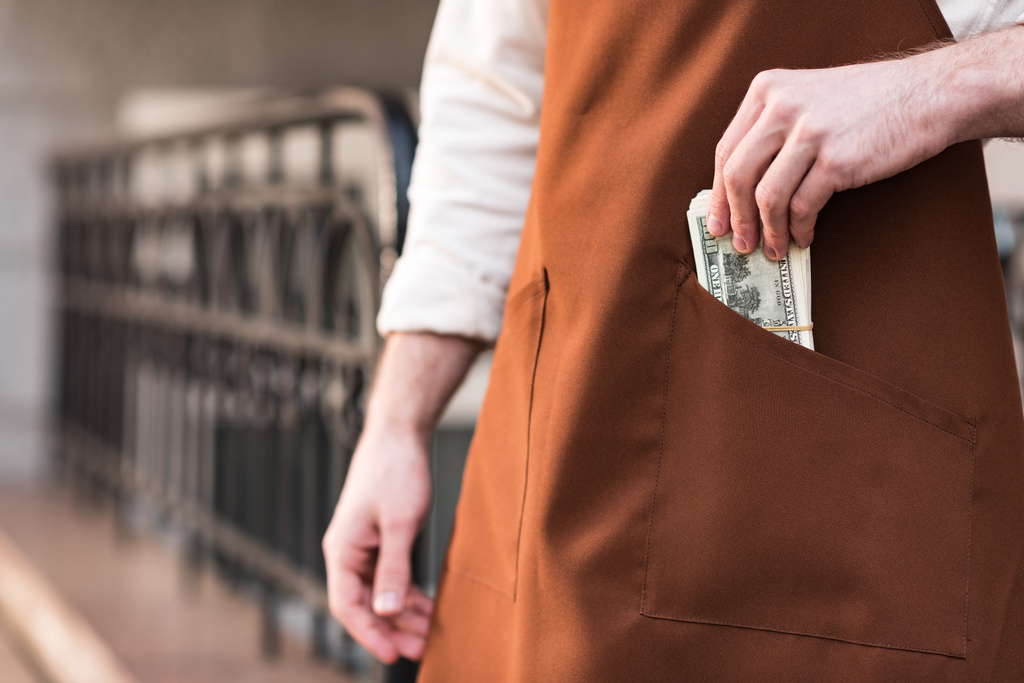 Частичный вид баристы в коричневом фартуке кладет деньги в карман
 - Фото, изображение