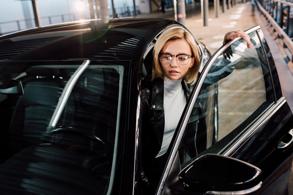 attraktives Mädchen mit Brille neben schwarzem Auto auf Parkplatz  - Foto, Bild