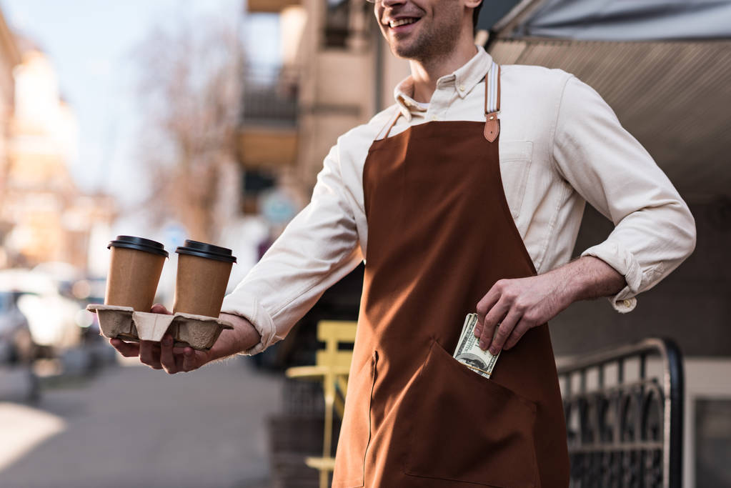 Vue recadrée du barista tenant des tasses en papier de café et mettant de l'argent dans sa poche
 - Photo, image
