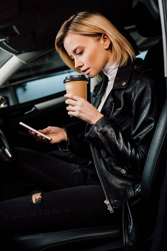 blondi nainen tilalla kertakäyttöinen kuppi ja käyttämällä älypuhelinta autossa
  - Valokuva, kuva