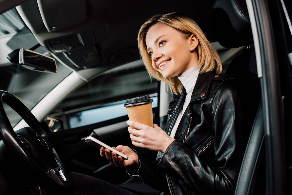 matala kulma näkymä iloinen blondi tyttö tilalla paperi kuppi ja älypuhelin autossa
  - Valokuva, kuva