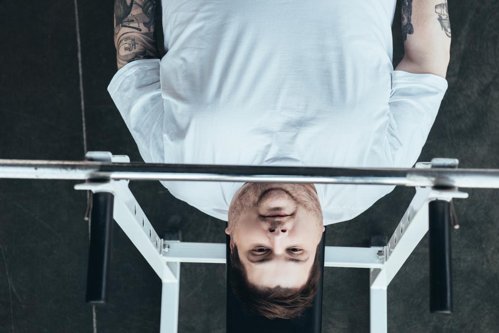 вид сверху на человека с татуировкой с избыточным весом, смотрящего на камеру во время тренировки с штангой в спортзале
 - Фото, изображение
