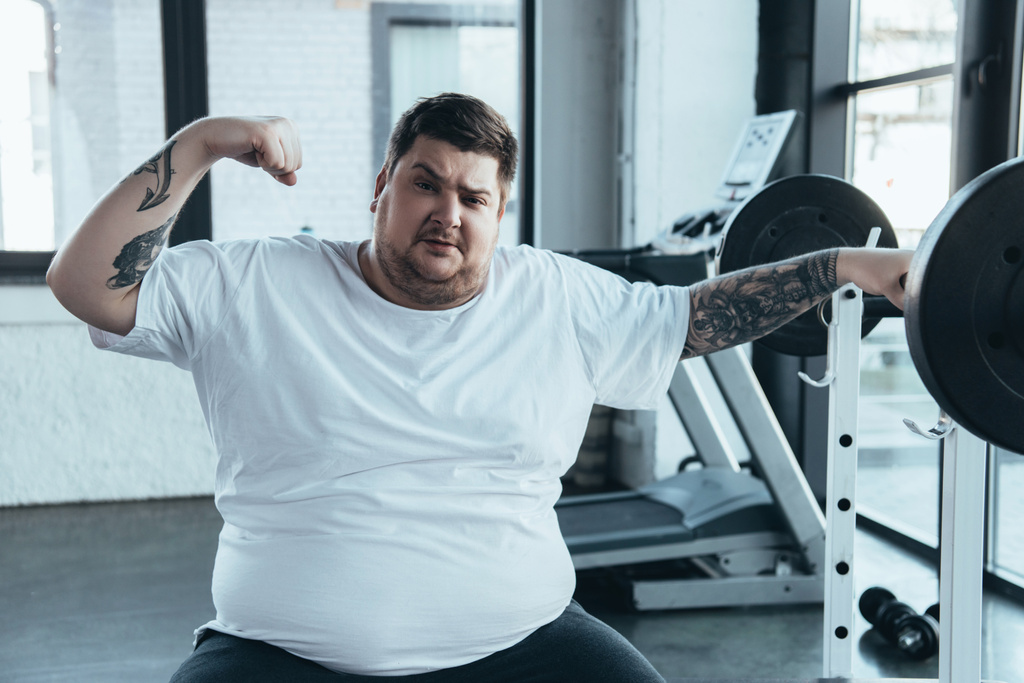 sobrepeso tatuado homem sentado no banco, olhando para a câmera e mostrando músculos no ginásio
 - Foto, Imagem