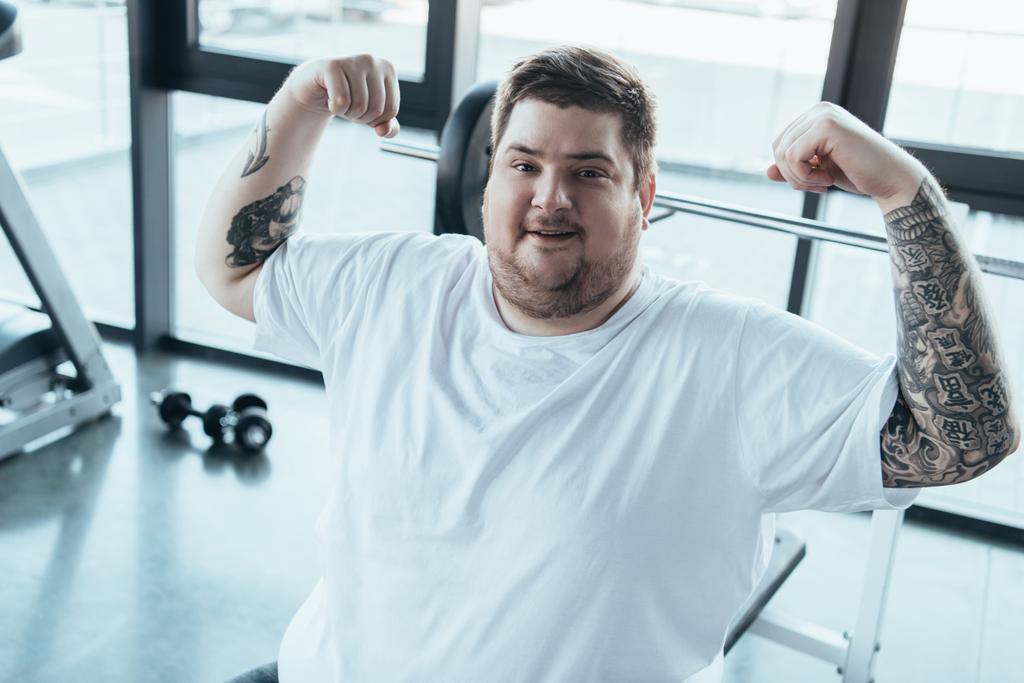 υπέρβαρο άντρας με τατουάζ χαμογελά, κοιτάζοντας την κάμερα και δείχνοντας τους μυς στο γυμναστήριο - Φωτογραφία, εικόνα