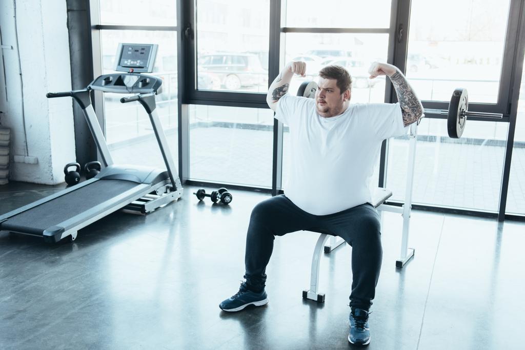 overweight tattooed man sitting on bench and showing muscles at gym - Photo, Image
