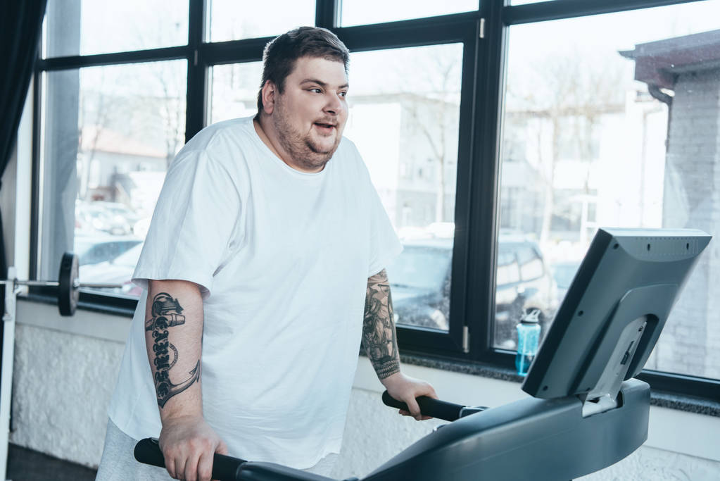 overweight tattooed man in white t-shirt running on treadmill at sports center - Photo, Image