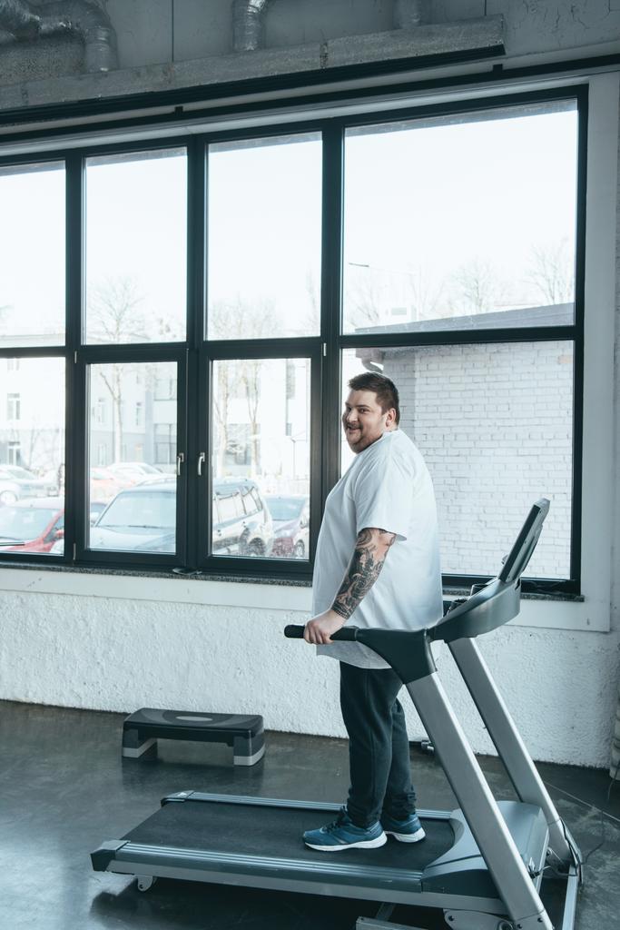 overweight tattooed man looking at camera while exercising on treadmill at sports center with copy space - Photo, Image