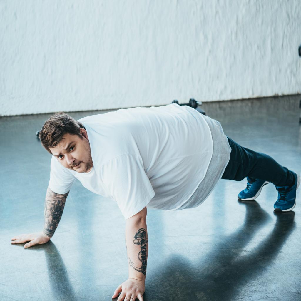 Tetovaný muž s nadváhou, který se dívá na kameru a přitom provádí cvičení ve sportovním centru - Fotografie, Obrázek