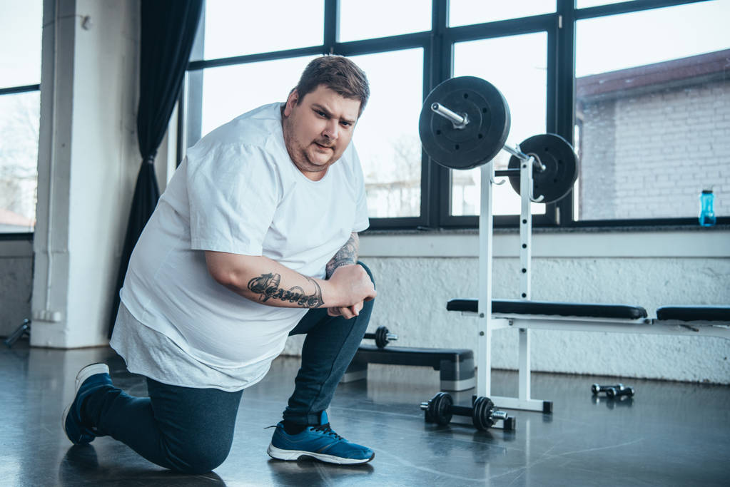 hombre tatuado con sobrepeso mirando la cámara y haciendo ejercicio de estiramiento en el gimnasio
 - Foto, imagen