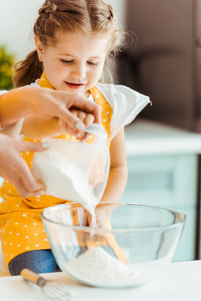 foco seletivo da mulher adicionando farinha para tigela perto da filha animada na cozinha
 - Foto, Imagem
