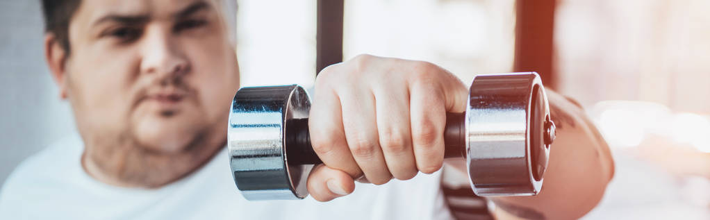 панорамный снимок человека с избыточным весом, смотрящего на камеру во время тренировки с гантелями в тренажерном зале
 - Фото, изображение