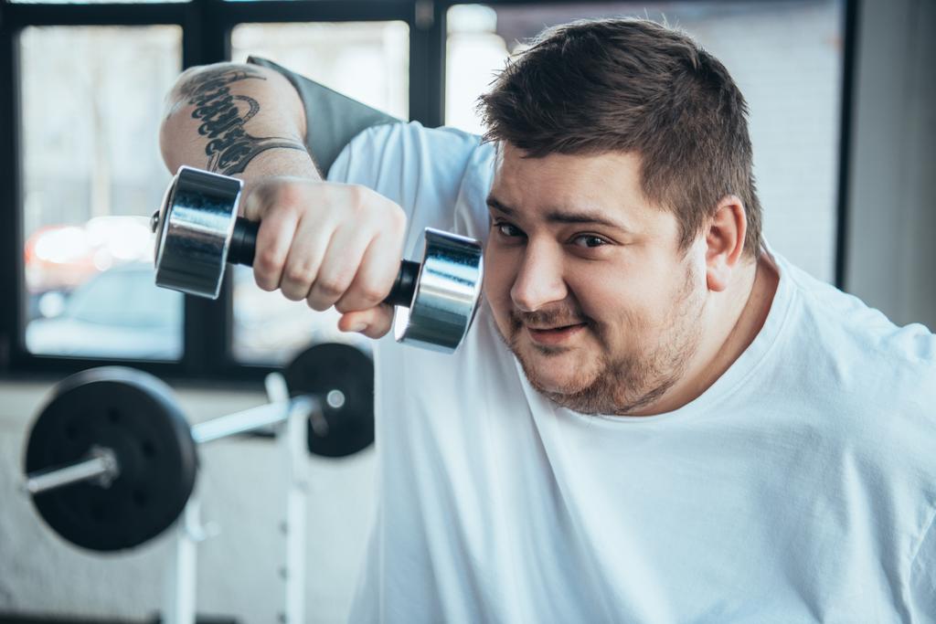 sorridente uomo tatuato sovrappeso Guardando la fotocamera e l'esercizio con i manubri al centro sportivo
 - Foto, immagini