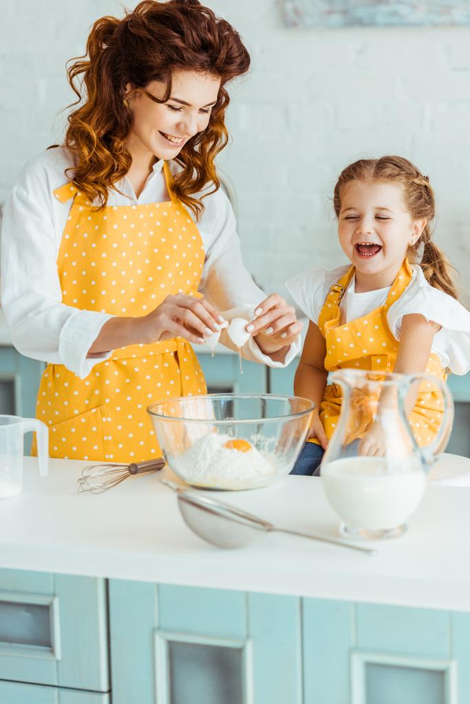 feliz madre e hija añadiendo huevo machacado a la harina en un tazón mientras cocinan juntas en la cocina
 - Foto, imagen
