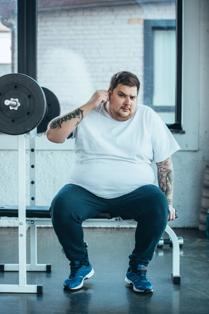 υπέρβαρο άντρας με τατουάζ στα ακουστικά που κάθονται στο παγκάκι, κοιτάζοντας την κάμερα και ακούγοντας μουσική στο γυμναστήριο - Φωτογραφία, εικόνα
