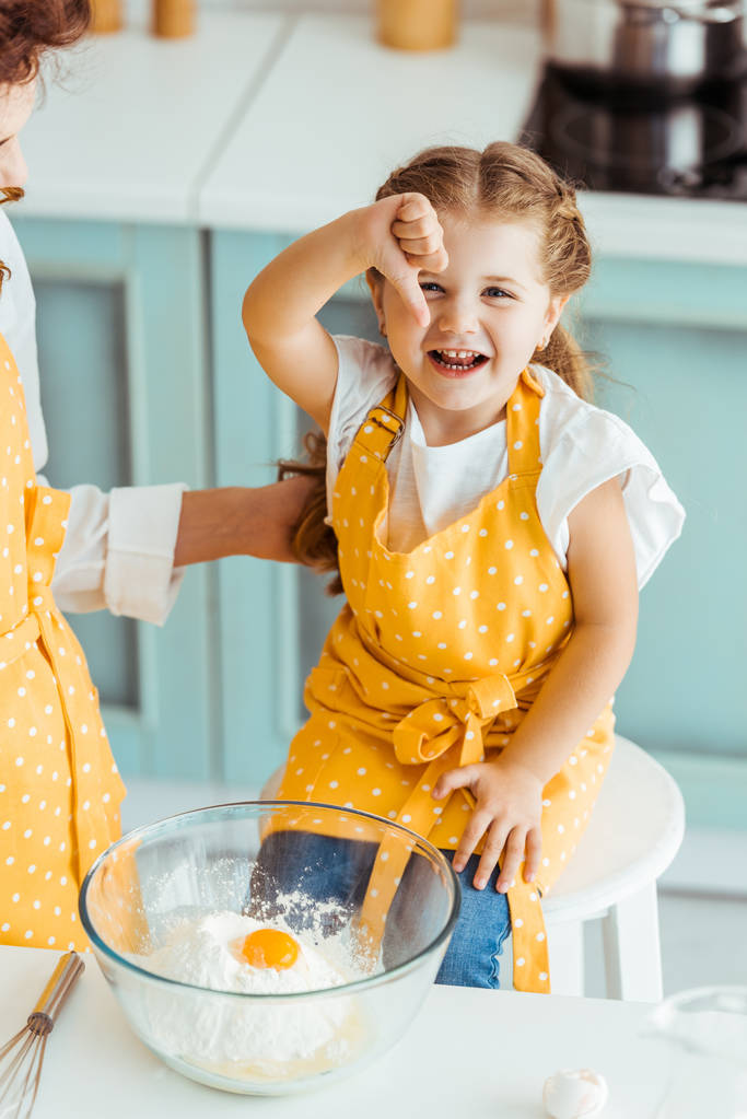 mother touching daughter in apron near bowl with flour and smashed egg while excited child showing thumb down - Photo, Image