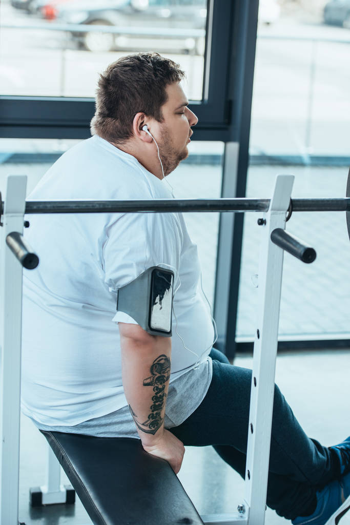 człowiek z nadwagą w słuchawkach siedzi na ławce i słuchania muzyki na siłowni - Zdjęcie, obraz