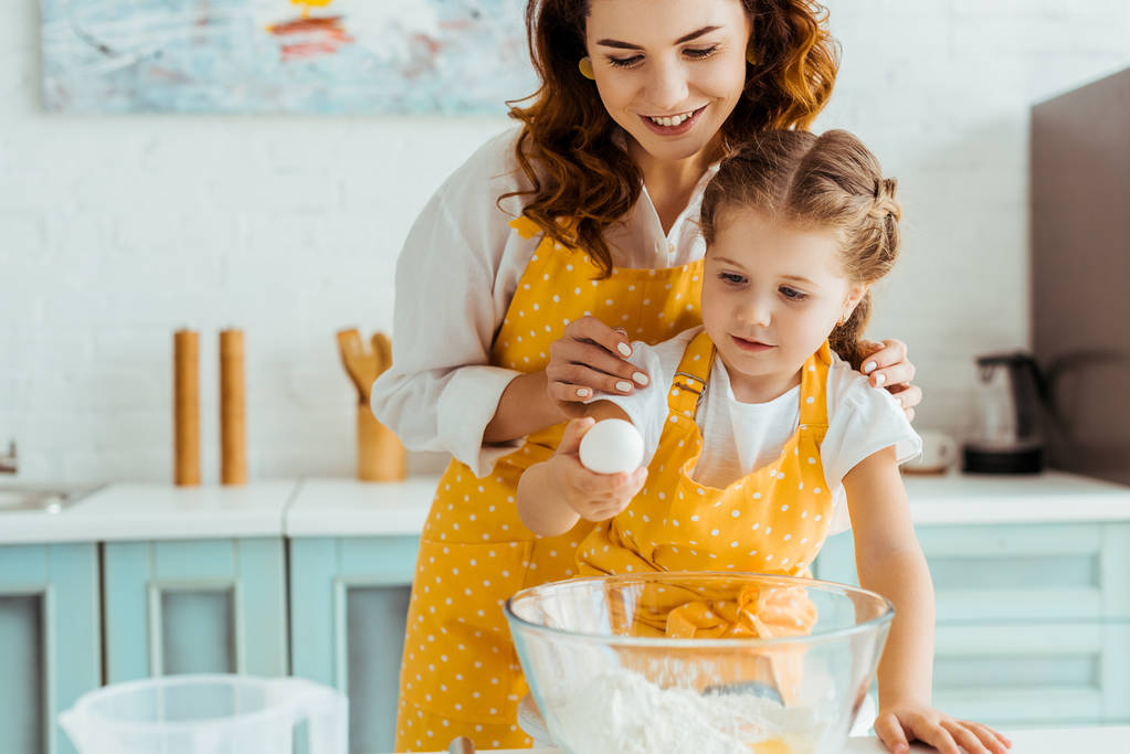 mãe e filha em aventais de bolinhas amarelas segurando ovo perto da tigela com farinha na cozinha
 - Foto, Imagem