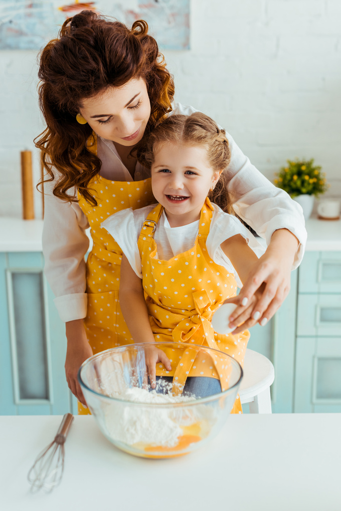 mother and excited daughter in yellow polka dot aprons near bowl with flour and eggs on table - Photo, Image
