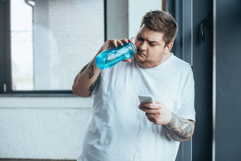 υπέρβαρο άντρας με τατουάζ χρησιμοποιώντας smartphone και πόσιμο νερό από το αθλητικό μπουκάλι στο γυμναστήριο - Φωτογραφία, εικόνα