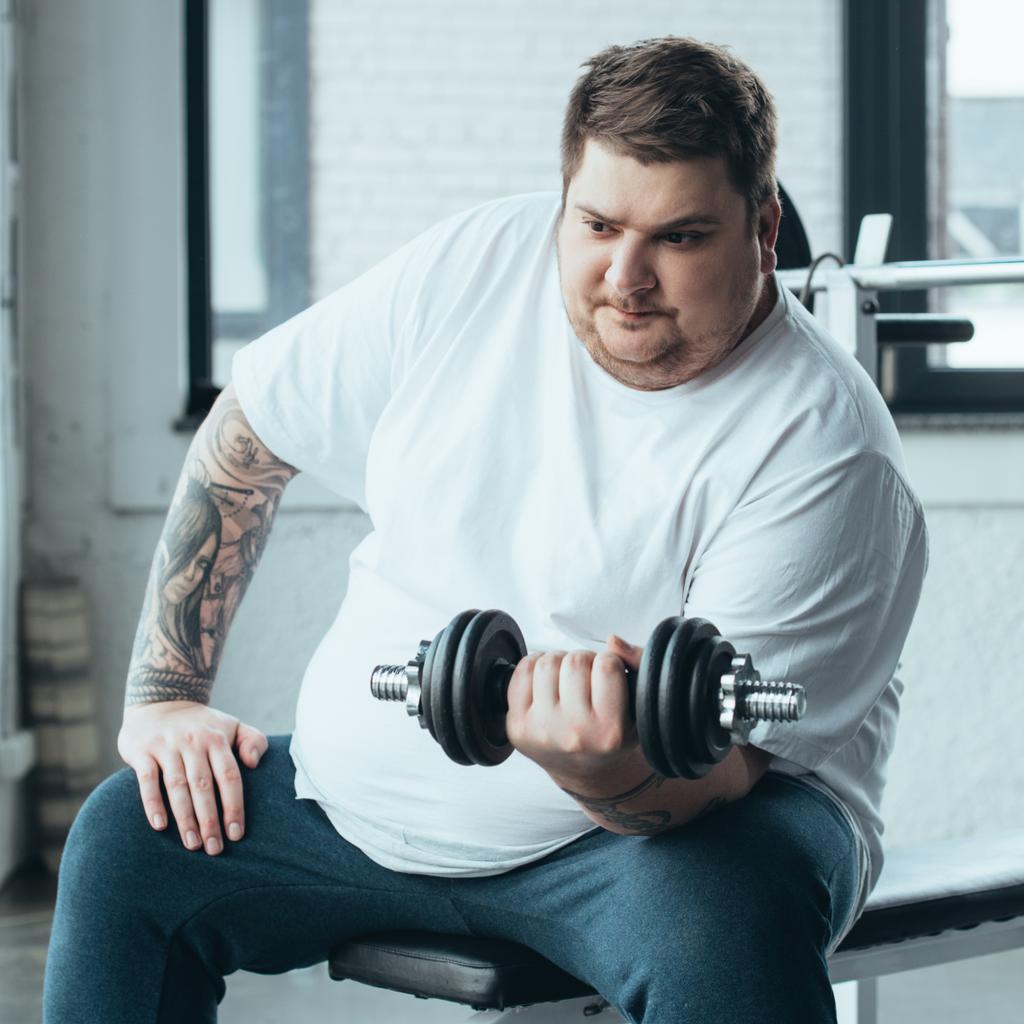 Толстый татуированный мужчина сидит и тренируется с гантелями в спорткомплексе
 - Фото, изображение
