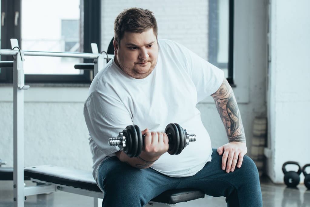 Overweight tattooed man sitting and training with dumbbell at sports center - Photo, Image