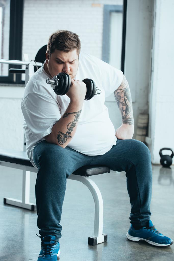 Sobrepeso tatuado homem sentado e treinando com haltere no centro de esportes
 - Foto, Imagem