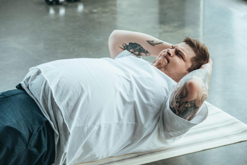 толстый татуированный мужчина лежит на коврике и делает сидя упражнения в спортивном центре
 - Фото, изображение