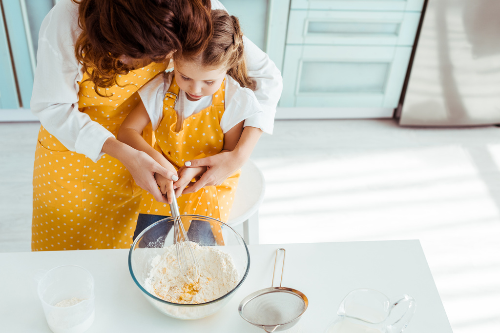 vista aerea di madre e figlia in grembiuli a pois mescolando farina e uova con frusta palloncino in ciotola
 - Foto, immagini