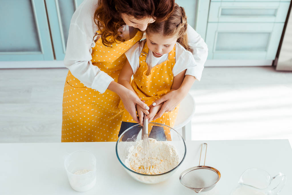 vista aérea de la madre y la hija en delantales mezclando harina y huevos con batidor de globo en un tazón
 - Foto, imagen