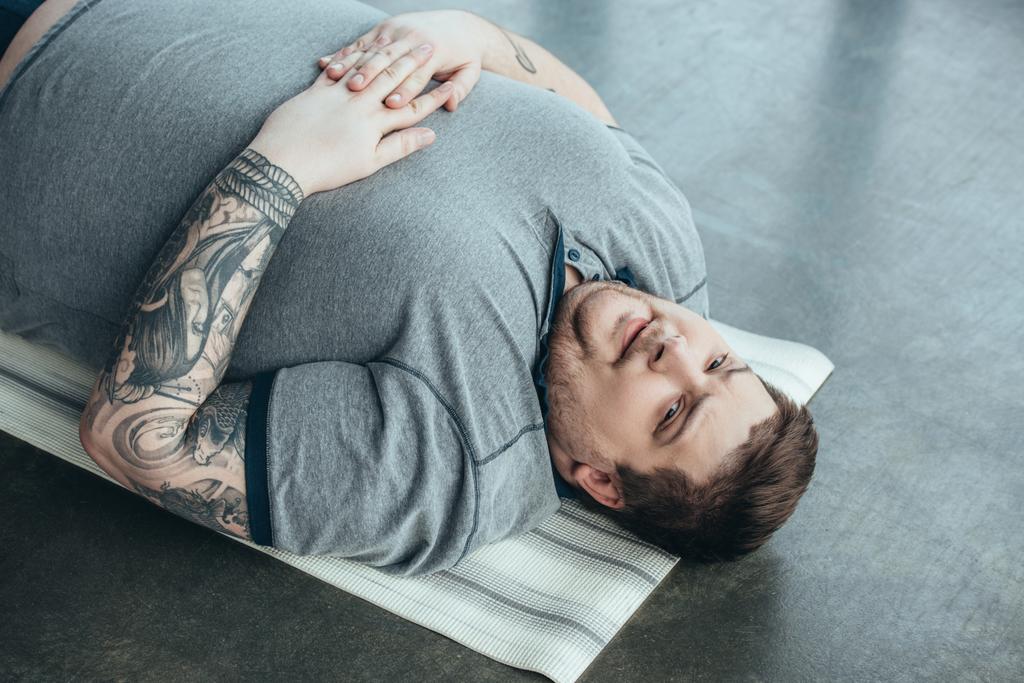 втомлений татуйований чоловік з надмірною вагою у сірій футболці, дивлячись на камеру, лежачи на фітнес-матусі у спортивному центрі
 - Фото, зображення