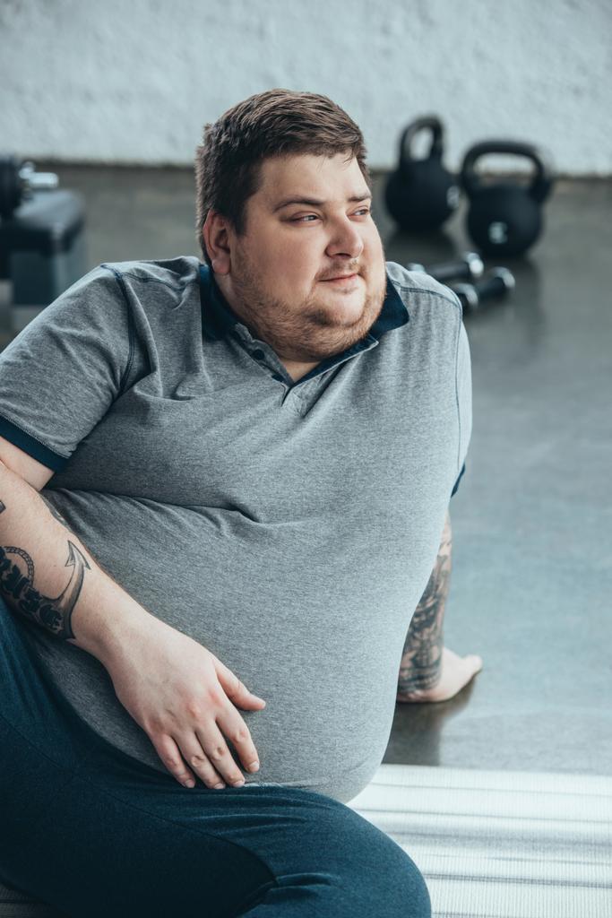 hombre tatuado con sobrepeso mirando hacia otro lado mientras está sentado en la colchoneta de fitness en el centro deportivo
 - Foto, imagen