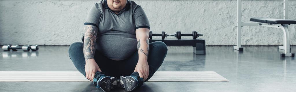Panoramaaufnahme eines übergewichtigen Mannes, der im Sportzentrum auf einer Fitnessmatte sitzt und die Beine streckt - Foto, Bild