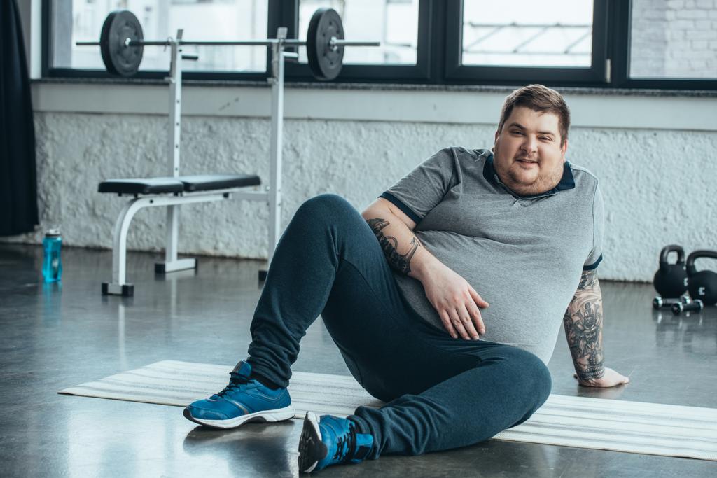 υπέρβαρα τατουάζ άνθρωπος κοιτάζοντας την κάμερα και κάθεται στο χαλάκι Fitness στο αθλητικό κέντρο - Φωτογραφία, εικόνα