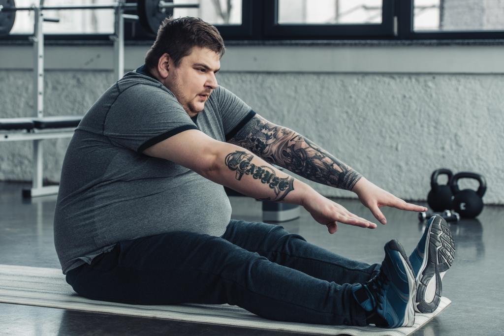 υπέρβαρων τατουάζ άνθρωπος κάθεται στο χαλάκι fitness και τέντωμα στο γυμναστήριο - Φωτογραφία, εικόνα