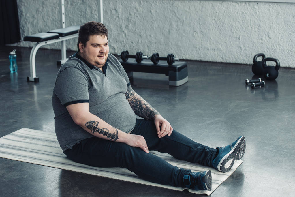 υπέρβαρο άντρας με τατουάζ σε γκρίζο μπλουζάκι που κάθεται στο χαλάκι Fitness στο αθλητικό κέντρο - Φωτογραφία, εικόνα