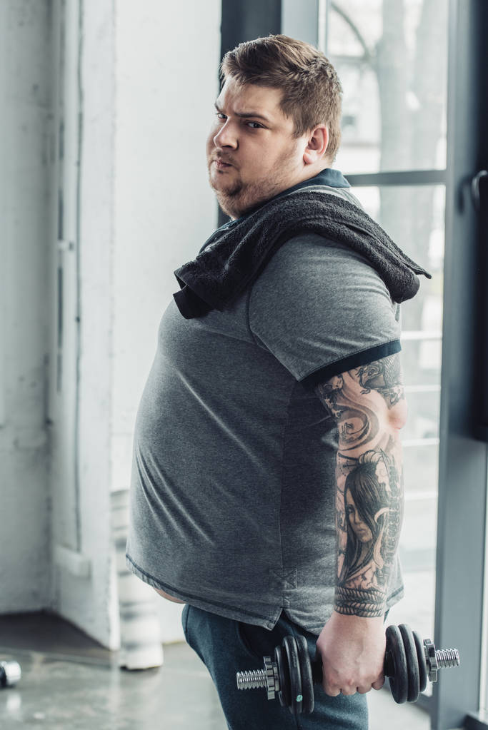 Obeso tatuato uomo guardando la fotocamera mentre si esercita con manubri presso il centro sportivo
 - Foto, immagini
