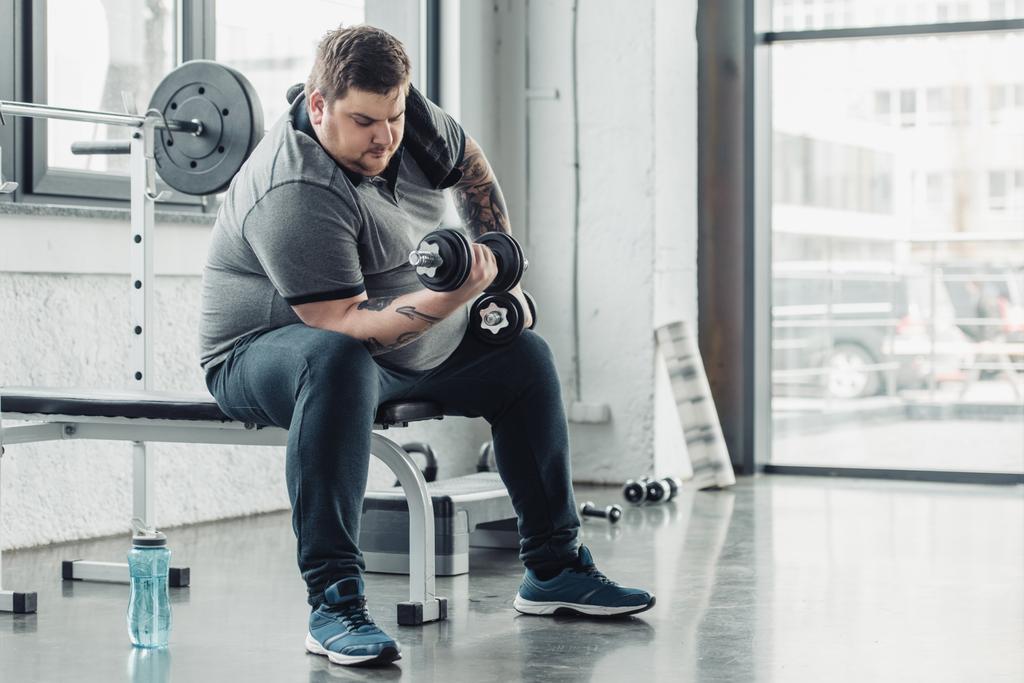 Übergewichtiger tätowierter Mann trainiert mit Kurzhanteln im Fitnessstudio mit Kopierraum - Foto, Bild