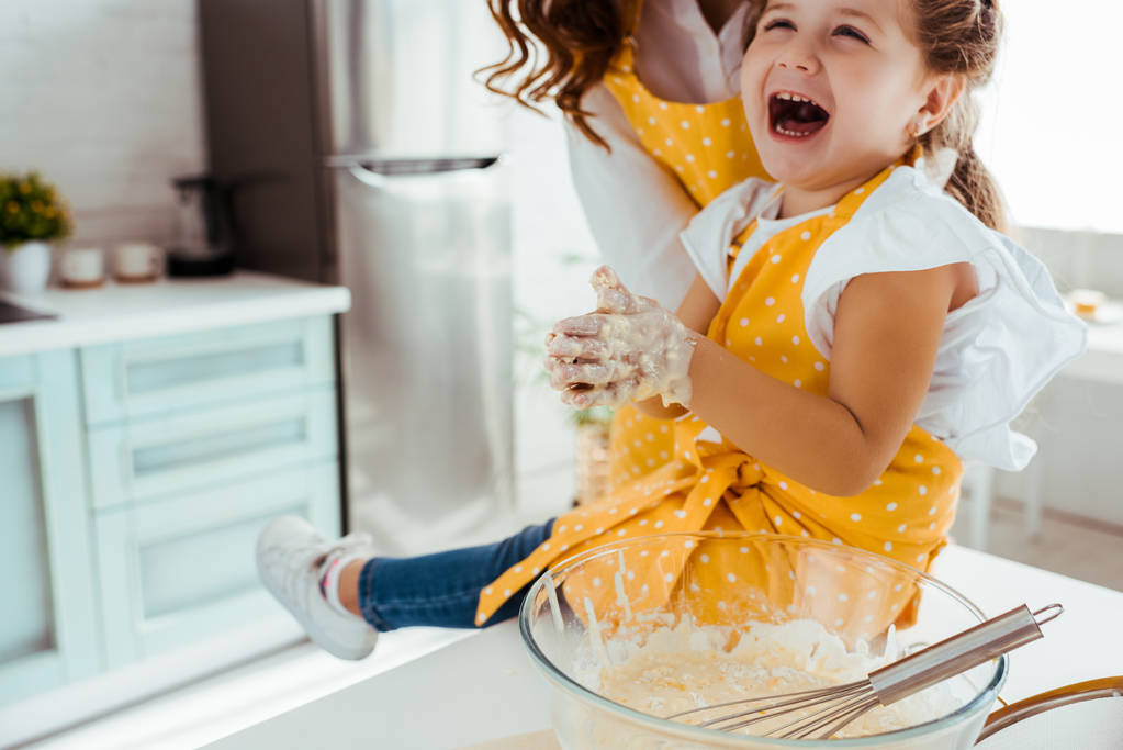 niño emocionado en delantal de lunares con masa en las manos riendo mientras está sentado junto a la madre en la mesa de la cocina
 - Foto, imagen