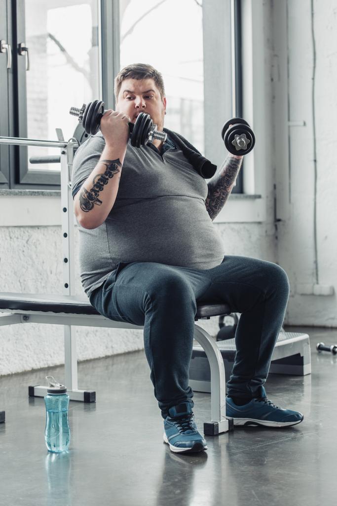 Избыточный вес татуированный мужчина сидит и тренируется с гантелями в спортивном центре
 - Фото, изображение