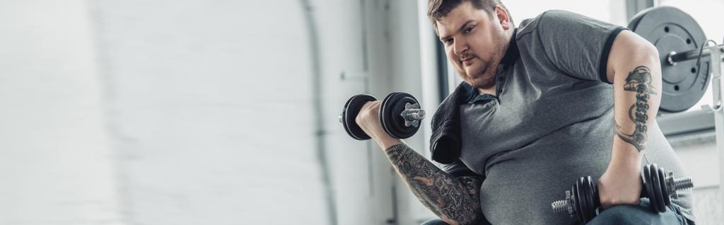 panoramic shot of Overweight tattooed man training with dumbbells at sports center - Photo, Image