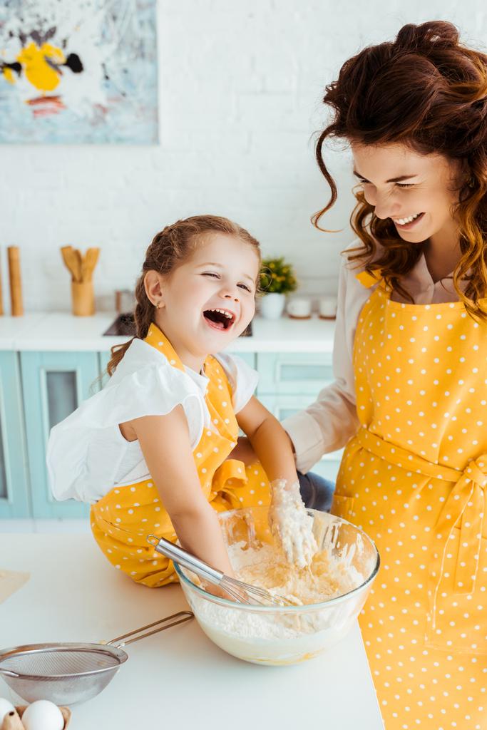 Улыбающаяся мать смотрит на смеющуюся счастливую дочь с руками в миске с тестом
 - Фото, изображение