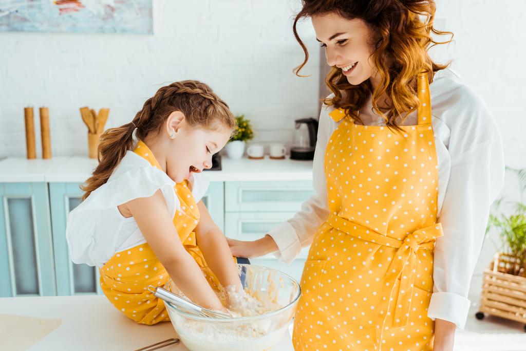 Lächelnde Mutter schaut glückliche Tochter an, die Hände in Schüssel mit Teig legt - Foto, Bild